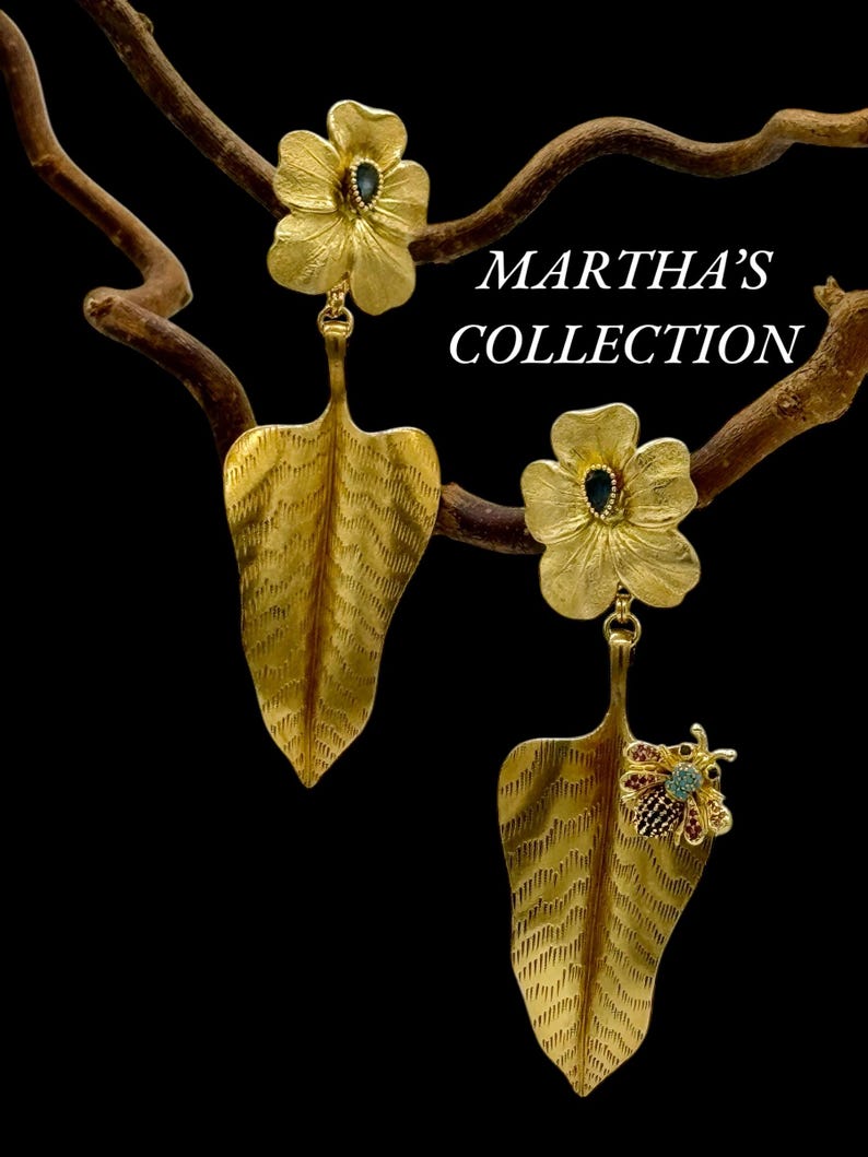 May include: Gold leaf earrings with a flower design and a small bee charm. The flowers have a blue gemstone in the centre. The earrings are hanging from a branch.