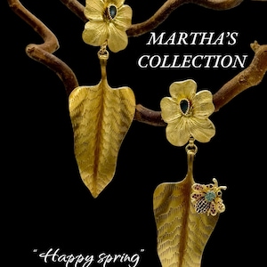 May include: Gold earrings featuring a flower design with a leaf dangle. The flower has a blue gemstone in the centre. The leaf has a textured surface and a small bee with red and blue gemstones on the bottom. The earrings are labelled "Martha's Collection" and "Happy Spring".