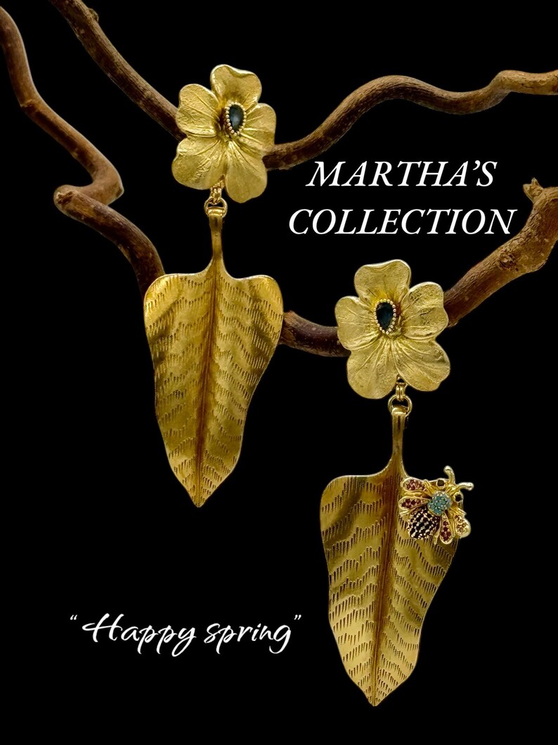 May include: Gold earrings featuring a flower design with a leaf dangle. The flower has a blue gemstone in the centre. The leaf has a textured surface and a small bee with red and blue gemstones on the bottom. The earrings are labelled "Martha's Collection" and "Happy Spring".