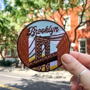 May include: A round embroidered patch with a brown and white image of the Brooklyn Bridge and the word "Brooklyn" in brown cursive lettering. The patch is held in a hand against a blurry background of a brick building and trees.