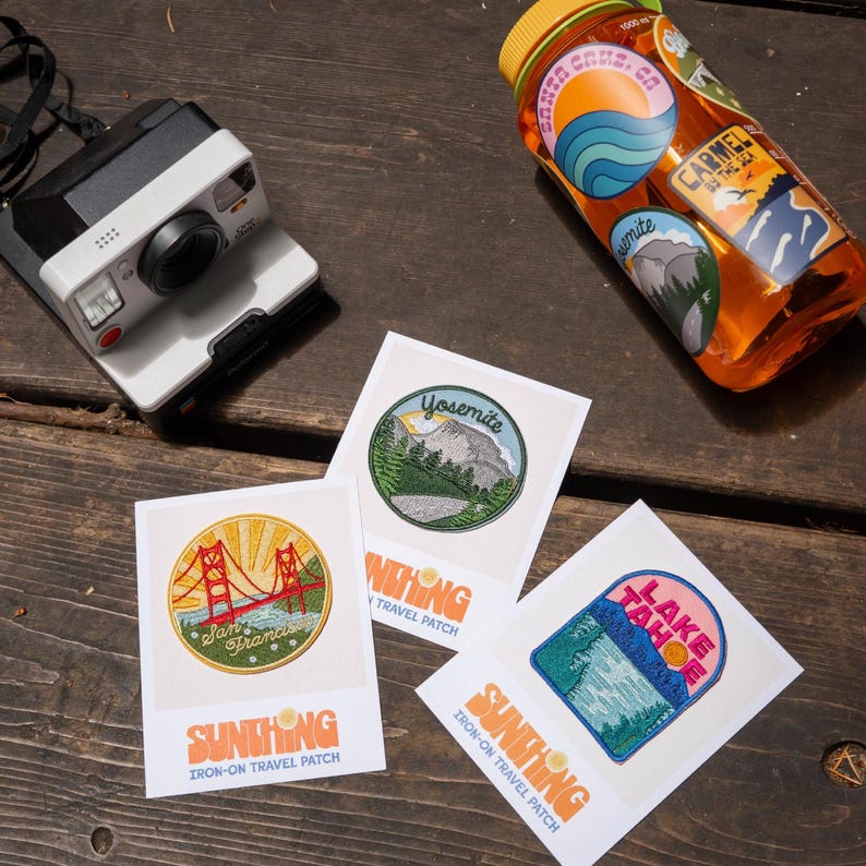 May include: Four iron-on travel patches featuring scenic landscapes and landmarks. The patches are on white cards with the text "Sunthing Iron-on Travel Patch". The patches feature Yosemite, San Francisco, Lake Tahoe, and Carmel by the Sea.