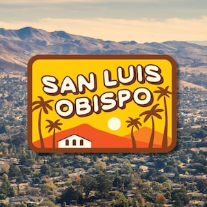 May include: A rectangular sticker with the text "SAN LUIS OBISPO" in white letters on a yellow background. The design includes palm trees, a white sun, and a red mountain range. The background shows a scenic view of a town and mountains.