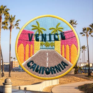 May include: A round, embroidered patch with a yellow border. The patch features a stylized image of the Venice Beach sign with palm trees and a road leading to it. The text "Venice California" is written in a green font.