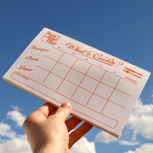 May include: A white weekly meal plan notepad with orange text and grid lines. The notepad includes the text "What's Cookin'?" and spaces for breakfast, lunch, and dinner. The notepad is held against a blue sky background.