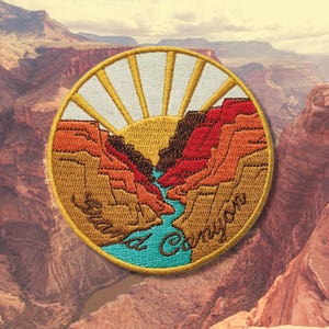 May include: Embroidered patch featuring a Grand Canyon landscape with a rising sun. The patch has a circular shape with a gold border. The design includes the text "Grand Canyon" in a cursive font, with shades of orange, brown, and blue.