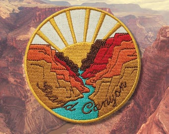 May include: Embroidered patch featuring a Grand Canyon landscape with a rising sun. The patch has a circular shape with a gold border. The design includes the text "Grand Canyon" in a cursive font, with shades of orange, brown, and blue.