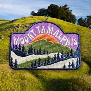 May include: Purple embroidered patch with the text "Mount Tamalpais" in white. The patch features a green mountain with an orange sunset behind it and a white winding road.