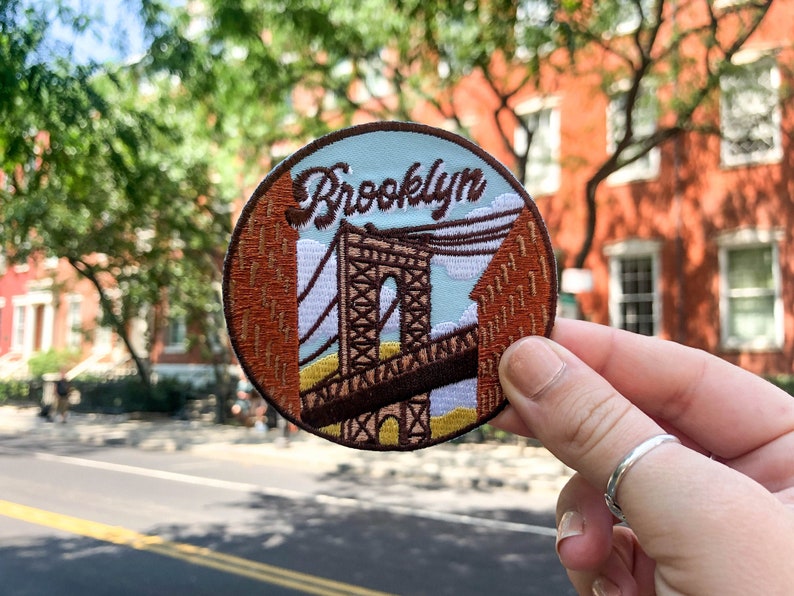 May include: A round embroidered patch with a brown and white image of the Brooklyn Bridge and the word "Brooklyn" in brown cursive lettering. The patch is held in a hand against a blurry background of a brick building and trees.