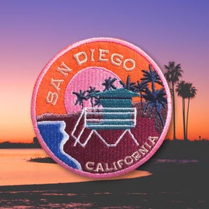 May include: Embroidered patch featuring a sunset scene with palm trees and a lifeguard tower. The patch is round with the words "San Diego California" in an orange and pink color scheme. The patch has a blue ocean and a pink sun.