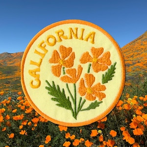 May include: Embroidered patch featuring orange California poppies and the word "CALIFORNIA" in yellow. The patch has a yellow background with an orange border. The background shows a field of orange poppies under a blue sky.