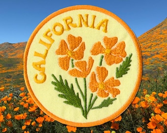 May include: Embroidered patch featuring orange California poppies and the word "CALIFORNIA" in yellow. The patch has a yellow background with an orange border. The background shows a field of orange poppies under a blue sky.