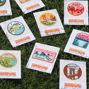 May include: Assortment of iron-on travel patches featuring various destinations, including Montauk, New York, San Francisco, Joshua Tree National Park, Lake Tahoe, Big Sur, California, Palm Springs, and Brooklyn. Each patch has a unique design and includes the text "SUNTHING IRON-ON TRAVEL PATCH".