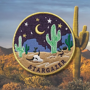 May include: Embroidered patch with a desert night scene. The patch features a crescent moon, stars, and cacti against a purple sky. The text "STARGAZER" is embroidered in gold around the bottom of the patch.