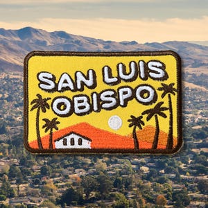 May include: A rectangular patch with the text "SAN LUIS OBISPO" in white letters. The patch features a yellow background with a landscape design of a mountain, palm trees, and a white building. The patch has a brown border.