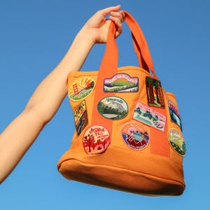 May include: Orange canvas tote bag with various embroidered patches featuring California landmarks and locations, including Yosemite, Mount Tamalpais, Joshua Tree National Park, and San Diego.