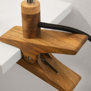 May include: A wooden clamp lamp with a black cord. The clamp is made of dark wood and has a metal screw. The lamp is attached to a white surface.