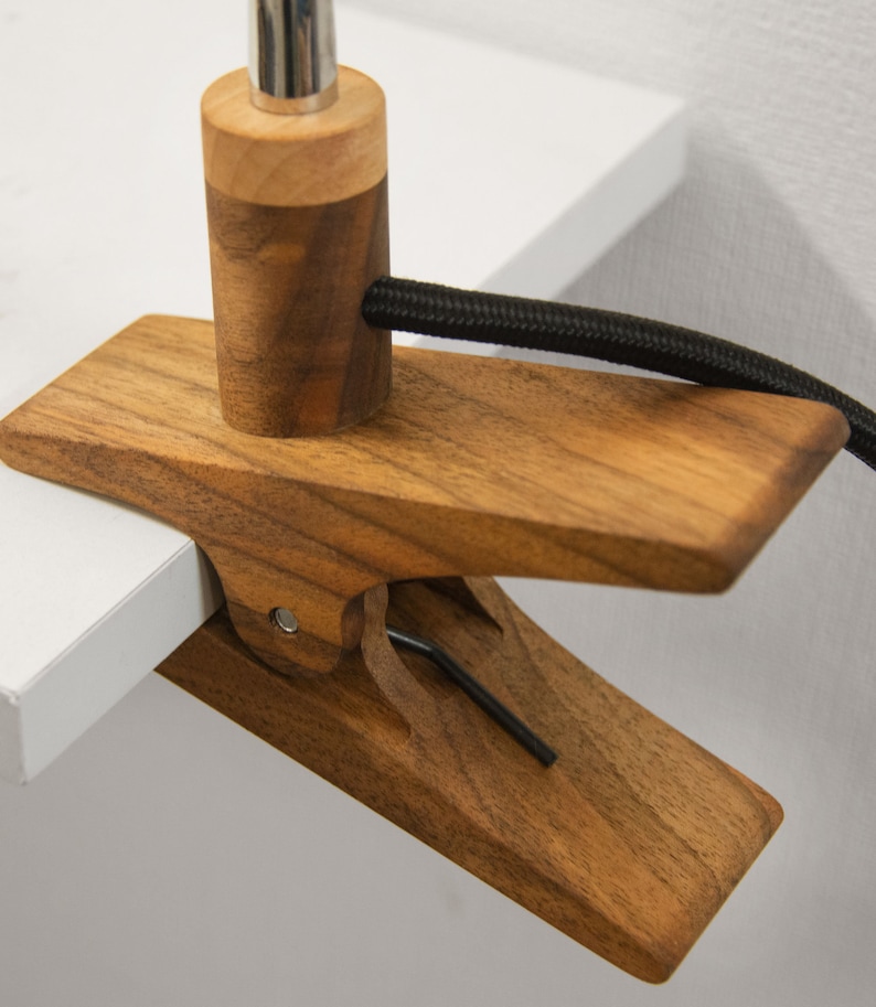 May include: A wooden clamp lamp with a black cord. The clamp is made of dark wood and has a metal screw. The lamp is attached to a white surface.