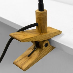 May include: A wooden desk lamp with a black cord and a clamp to attach to a desk or table. The lamp has a flexible arm and a black metal shade.