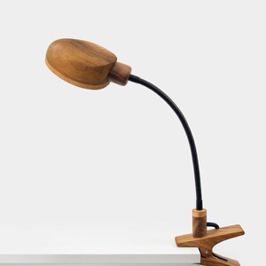 May include: A wooden desk lamp with a clamp base and a flexible gooseneck. The lamp has a natural wood finish and a round, oval-shaped shade.