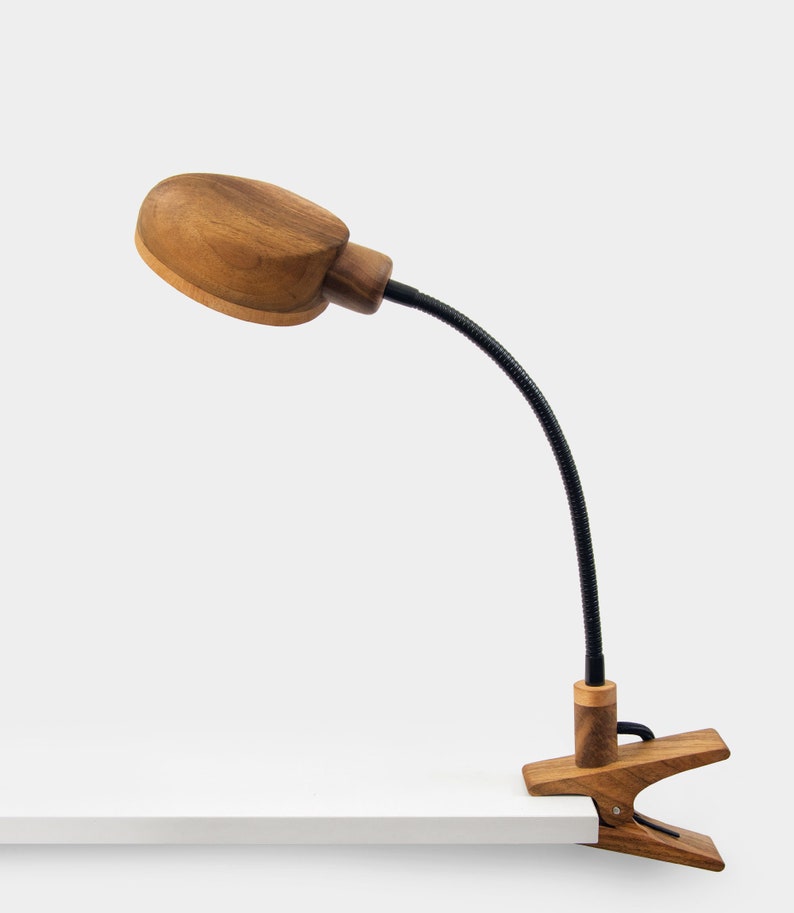 May include: A wooden desk lamp with a clamp base and a flexible gooseneck. The lamp has a natural wood finish and a round, oval-shaped shade.