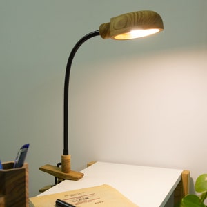 May include: A wooden desk lamp with a black flexible arm and a clamp. The lamp is on and illuminating a white desk surface.