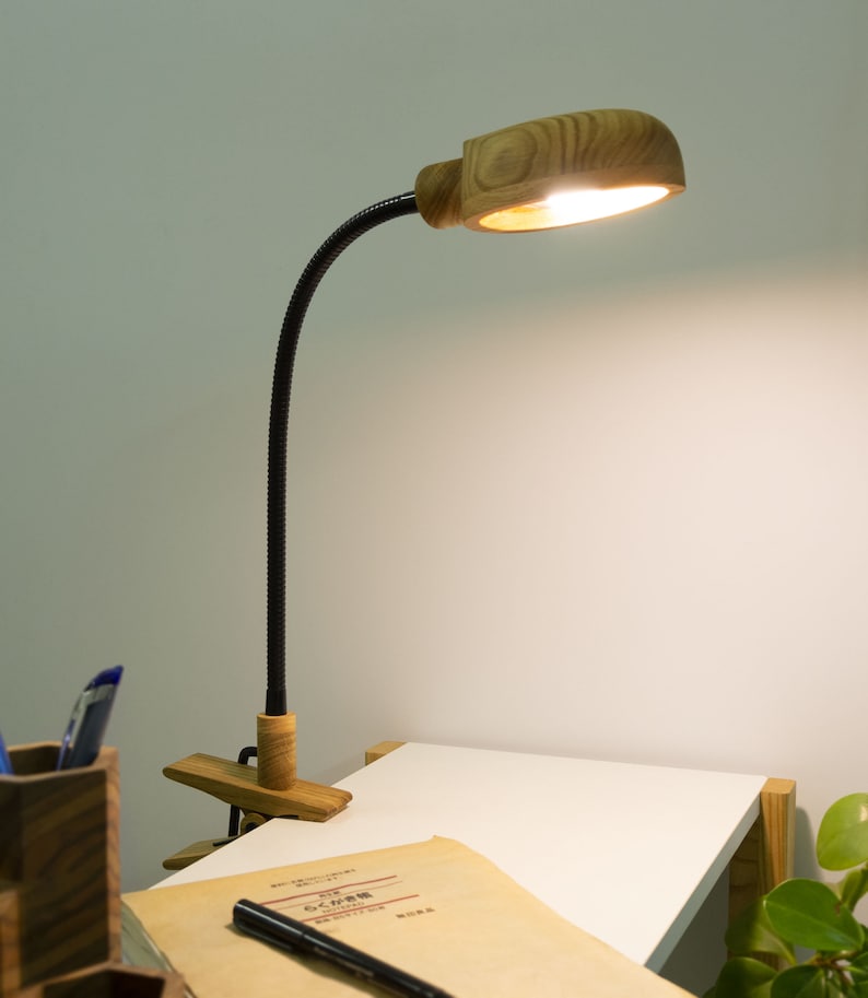 May include: A wooden desk lamp with a black flexible arm and a clamp. The lamp is on and illuminating a white desk surface.