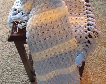May include: A blue and white crocheted baby blanket with fringe. The blanket is folded and draped over a wooden table.