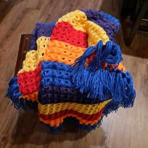 May include: A handmade, colorful crochet blanket. The blanket features a vibrant array of colors, including yellow, red, orange, blue, and purple. The edges are trimmed with blue fringe. The blanket is folded and appears to be made of soft yarn.