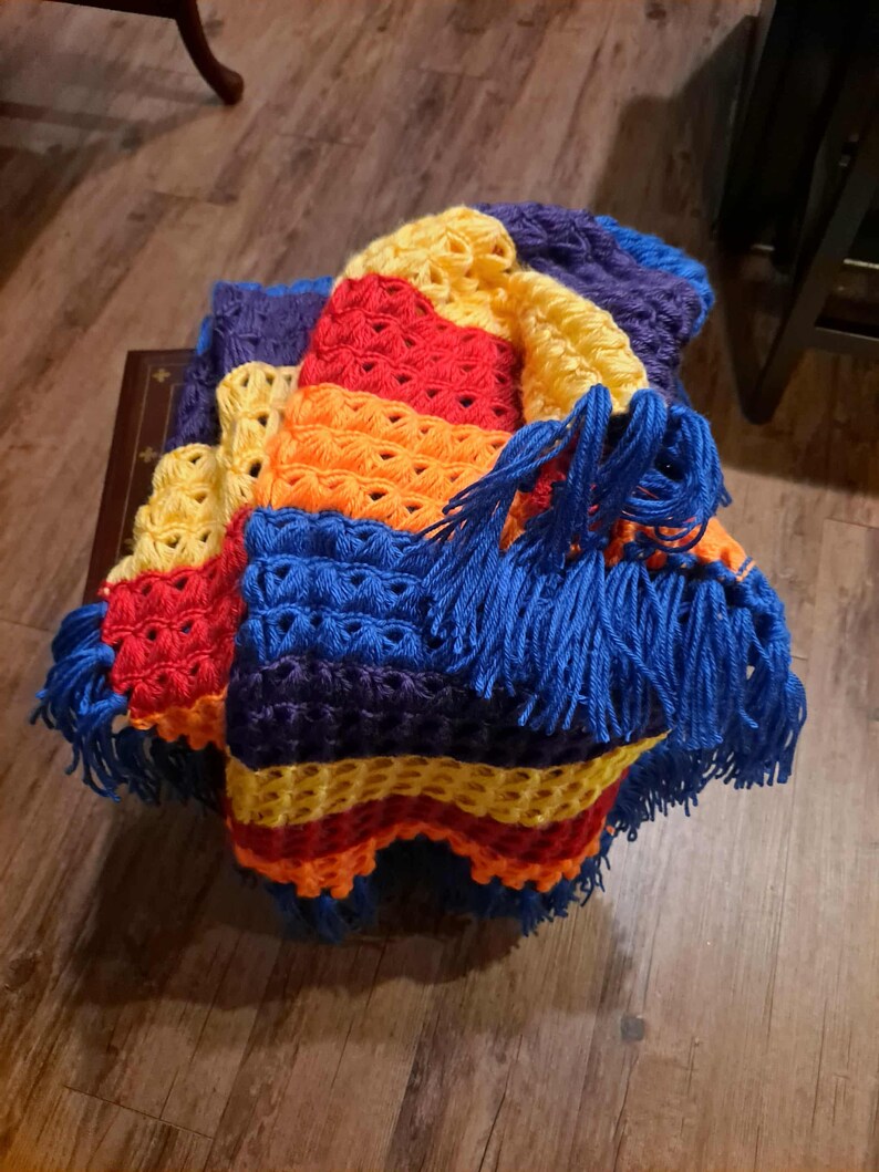 May include: A handmade, colorful crochet blanket. The blanket features a vibrant array of colors, including yellow, red, orange, blue, and purple. The edges are trimmed with blue fringe. The blanket is folded and appears to be made of soft yarn.
