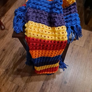 May include: A colorful, handmade crochet blanket with a vibrant rainbow pattern. The blanket features a combination of blue, purple, yellow, orange, and red hues. It has a textured design and is adorned with blue fringe.