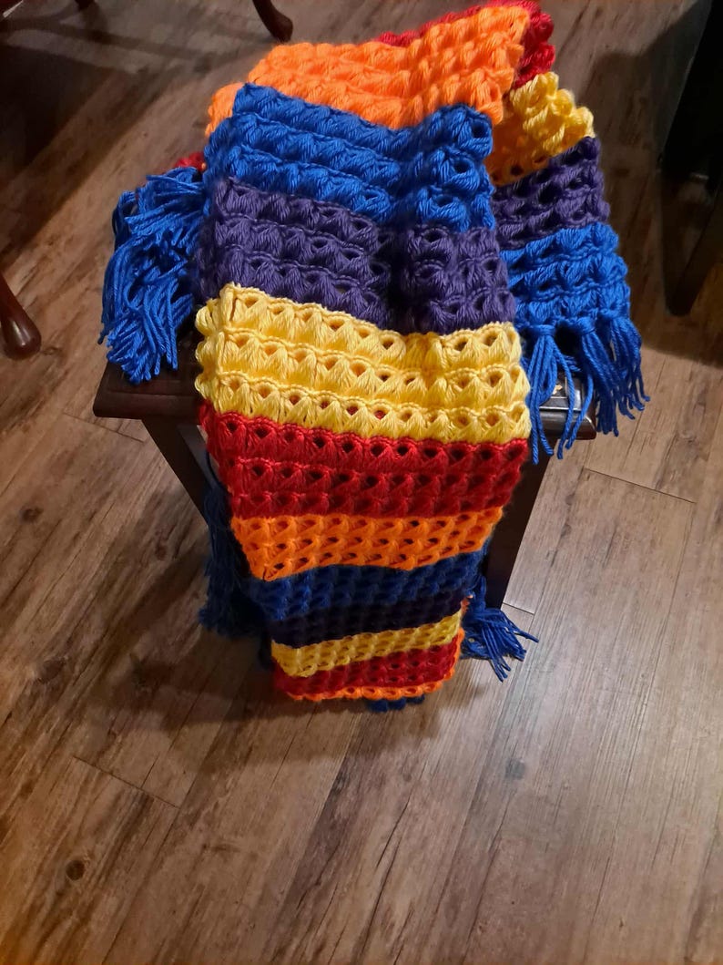 May include: A colorful, handmade crochet blanket with a vibrant rainbow pattern. The blanket features a combination of blue, purple, yellow, orange, and red hues. It has a textured design and is adorned with blue fringe.