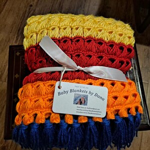 May include: A stack of handmade baby blankets in yellow, red, orange, and blue. The blankets are tied with a white ribbon and a tag that reads "Baby Blankets by Dana". The blankets are made in the USA.