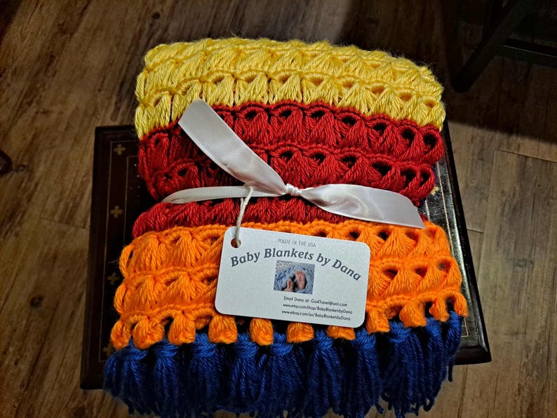 May include: A stack of handmade baby blankets in yellow, red, orange, and blue. The blankets are tied with a white ribbon and a tag that reads "Baby Blankets by Dana". The blankets are made in the USA.