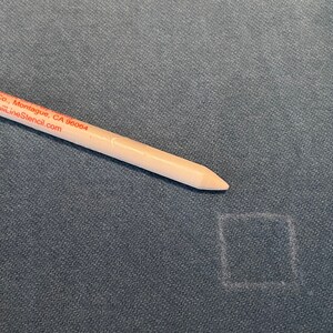 May include: A white marking tool with the text "Mfg. Co., Montague, CA 96064 www.FujiLineStencil.com" on it, is lying on a dark gray fabric surface. A white square is drawn on the fabric.