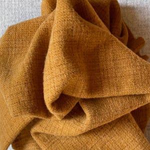 May include: Mustard-colored scarf made of a soft, textured material. The scarf is folded and draped, showcasing its warm, earthy tone. The fabric appears to be thick and cozy, perfect for cooler weather. The scarf is a solid color with no visible patterns.