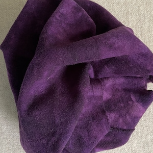 May include: A close-up of a deep purple fabric, possibly velvet or suede, with visible texture and folds. The fabric is bunched and draped, showcasing its rich color and soft appearance. The background is a neutral gray.