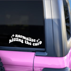 May include: A pink car with a white vinyl sticker on the back window that says "normalize hitting the curb" with stars on either side of the text.