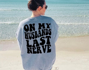 Puede incluir: Una sudadera gris con el texto "On My Husbands Last Nerve" impreso en negro en la espalda.