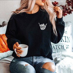 May include: Black crewneck sweatshirt with a white skeleton hand graphic on the chest. The sweatshirt is a solid black color and has long sleeves. The graphic is a stylized depiction of a hand making a 'hang loose' gesture.