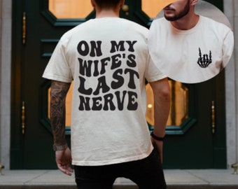 Puede incluir: Una camiseta blanca con el texto "On My Wife's Last Nerve" impreso en negro en la espalda. La parte delantera de la camiseta tiene una pequeña mano de esqueleto negro con un gesto de rock and roll.