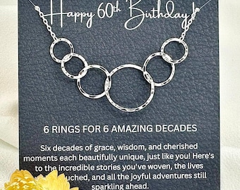May include: A silver necklace with six interlocking rings is displayed on a dark gray card. The card reads "Happy 60th Birthday!" and "6 Rings for 6 Amazing Decades." A yellow flower is in the foreground.