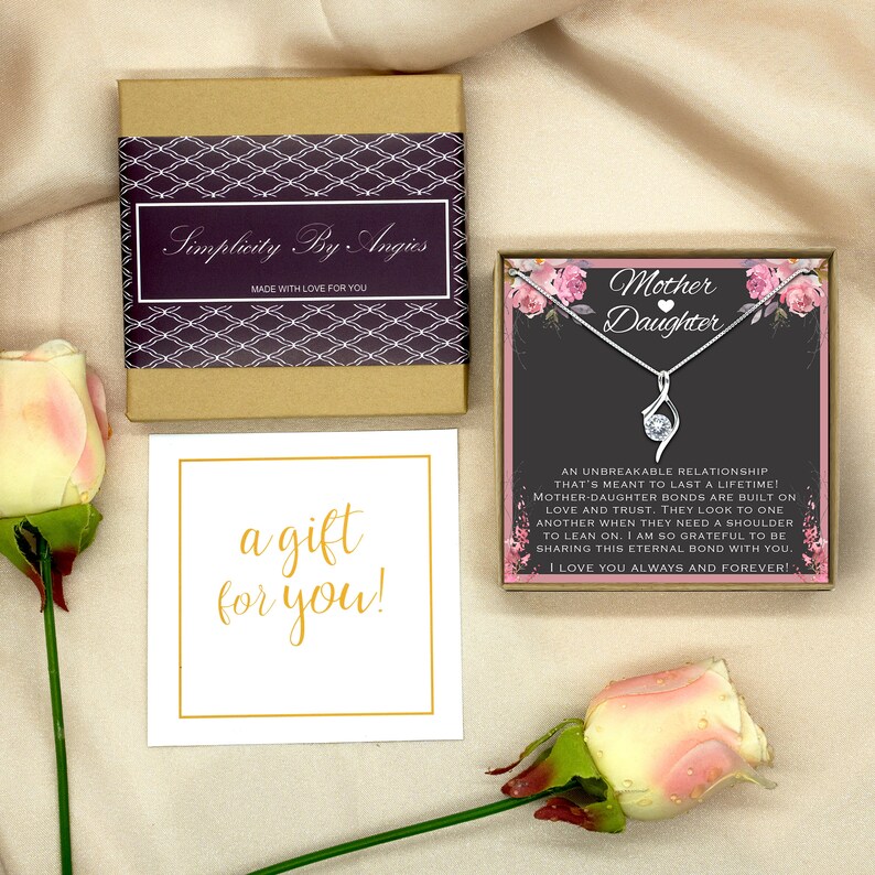 May include: A gift box with a white card that says "a gift for you!" and a small box with a silver necklace with a clear stone pendant. The box has a pink and white floral design and the text "Mother Daughter" with a heart symbol. The text on the box reads "An unbreakable relationship that's meant to last a lifetime! Mother daughter bonds are built on love and trust. They look to one another when they need a shoulder to lean on. I am so grateful to be sharing this eternal bond with you. I love you always and forever!"