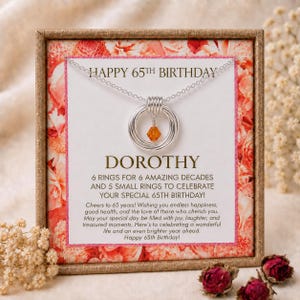 May include: A silver necklace with a pendant of six interlocking rings and a small orange crystal charm, presented on a card that reads HAPPY 65TH BIRTHDAY DOROTHY, 6 RINGS FOR 6 AMAZING DECADES AND 5 SMALL RINGS TO CELEBRATE YOUR SPECIAL 65TH BIRTHDAY.
