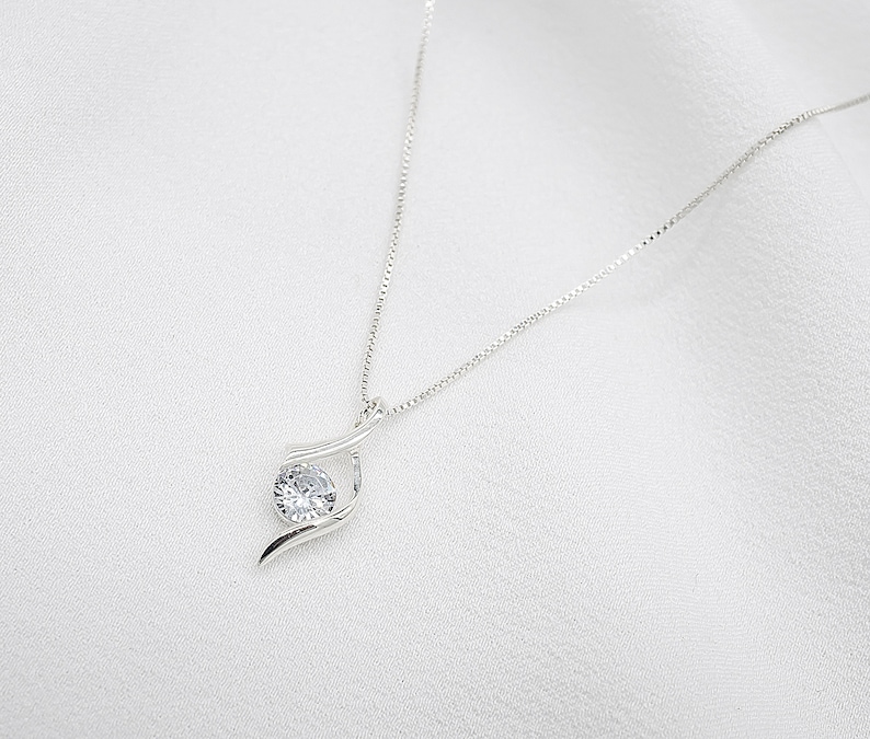 May include: A silver necklace with a delicate chain and a single, clear, round gemstone pendant. The pendant is set in a simple, elegant design.