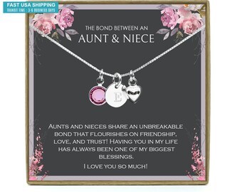 May include: A silver necklace with a heart charm, a charm with a pink crystal, and a charm with the letter "E". The necklace is in a box with a black background and pink floral accents. The text on the box reads "The bond between an aunt & niece" and "AUNTS AND NIECES SHARE AN UNBREAKABLE BOND THAT FLOURISHES ON FRIENDSHIP, LOVE, AND TRUST! HAVING YOU IN MY LIFE HAS ALWAYS BEEN ONE OF MY BIGGEST BLESSINGS. I LOVE YOU SO MUCH!"