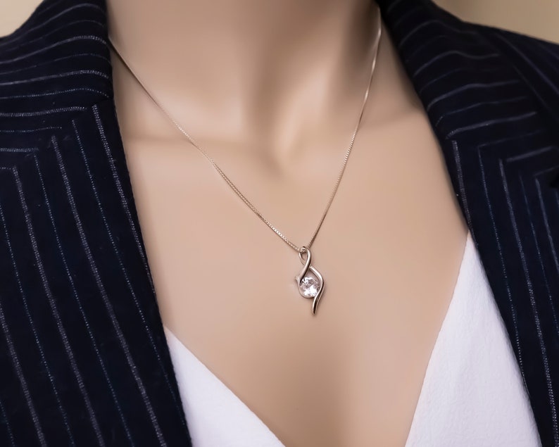 May include: A silver necklace with a delicate pendant featuring a clear gemstone. The pendant is shaped like a stylized infinity symbol.