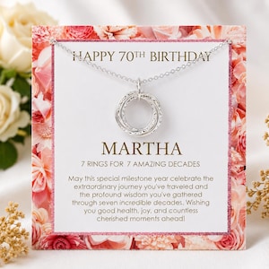 Peut inclure: Un collier en argent avec un pendentif composé de sept anneaux entrelacés sur une carte à bordure florale. Le texte indique : HAPPY 70TH BIRTHDAY, MARTHA, 7 RINGS FOR 7 AMAZING DECADES, ainsi qu'un message célébrant sept décennies de vie.