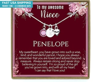 May include: A silver necklace with three charms, including a heart, a round charm with a pink stone, and a disc with the letter "E". The necklace is displayed on a card with the text "To my awesome Niece" and the name "PENELOPE".