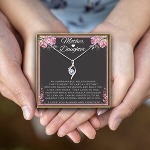 May include: A silver necklace with a delicate infinity pendant and a small clear crystal. The necklace is displayed in a gift box with a pink floral design and the words "Mother Daughter" on the front. The box is held by two hands.