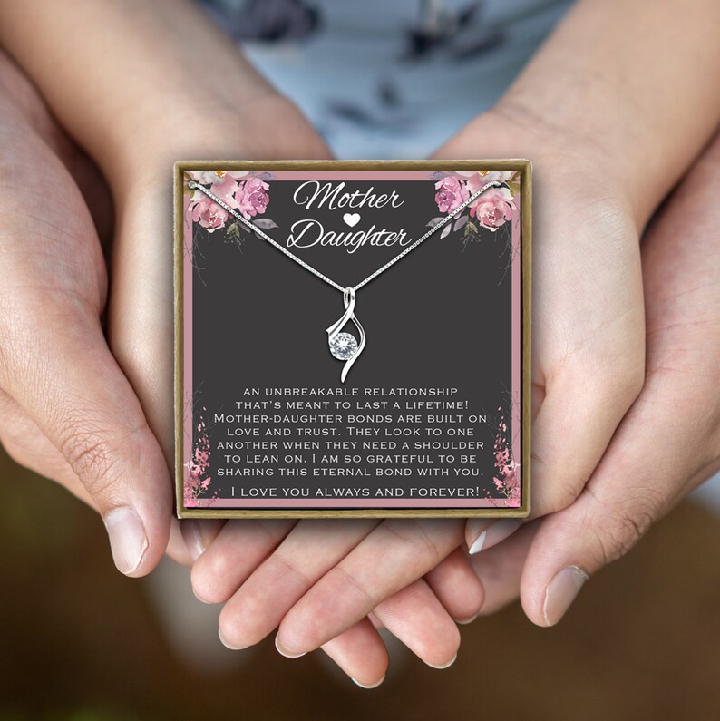 May include: A silver necklace with a delicate infinity pendant and a small clear crystal. The necklace is displayed in a gift box with a pink floral design and the words "Mother Daughter" on the front. The box is held by two hands.